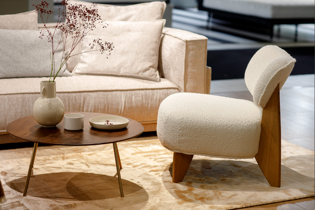 A white upholstered armchair with a wooden frame, featuring a contemporary design, placed in a room with a round wooden table and a beige sofa in the background.