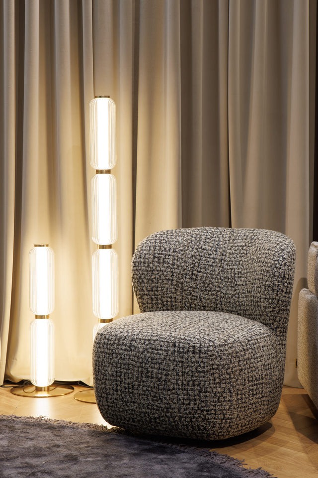 A gray upholstered armchair with a textured pattern, positioned in a room with a curtain backdrop, illuminated by a floor lamp.